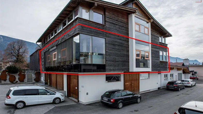 Modernes Geschäftsgebäude mit Holzfassade und großen Fensterfronten, umgeben von Parkplätzen und Bergkulisse.