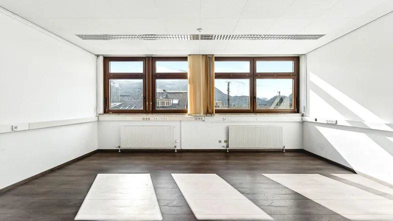 Heller, leerer Raum mit großen Fenstern, die viel Tageslicht hereinlassen und einen Blick nach draußen bieten.