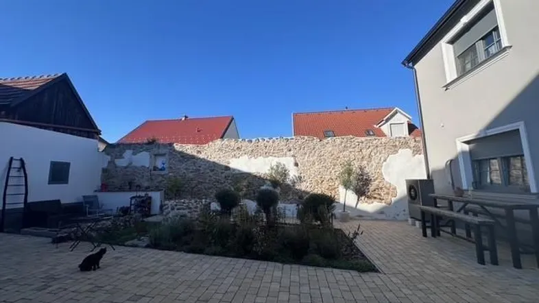 Gepflasterter Innenhof mit Steinmauer, Sitzgelegenheiten und kleiner Bepflanzung unter blauem Himmel.