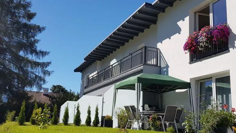 Außenansicht des Hauses mit Garten, möblierter Sitzecke und Balkon unter blauem Himmel.