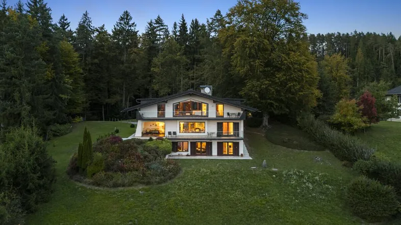 Elegantes Anwesen bei Dämmerung, beleuchtet und eingebettet in eine grüne Waldlandschaft.
