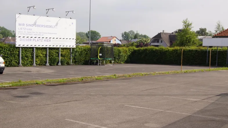 Großer asphaltierter Parkplatz mit Werbetafel und Hecke im Hintergrund, umgeben von Wohnhäusern.