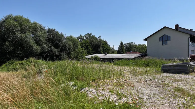 Unbebautes Grundstück mit Gras, Bäumen und einem Haus im Hintergrund unter blauem Himmel.