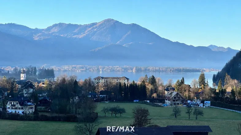 Panoramablick auf den Wolfgangsee mit umliegenden Bergen und einem Dorf mit Neubauprojekt.
