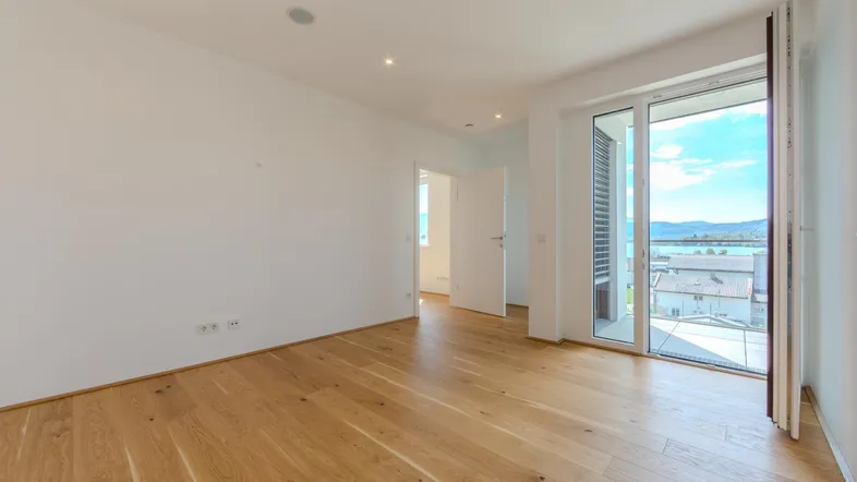 Helles Zimmer mit Holzboden und Zugang zu einem Balkon mit See- und Bergblick.