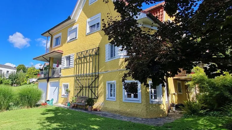 Gelbes Wohnhaus mit grünem Garten und blauer Himmel, umgeben von Bäumen und Sträuchern.