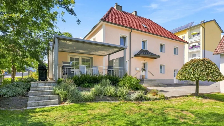 Hausfassade mit überdachter Terrasse, gepflegtem Garten und Zufahrt zum Grundstück.
