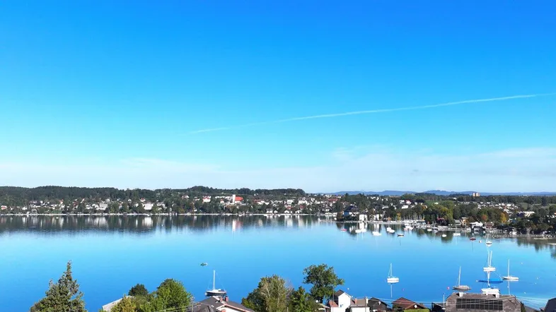 Panoramablick auf den See mit umliegenden Häusern und grünen Hügeln unter blauem Himmel.