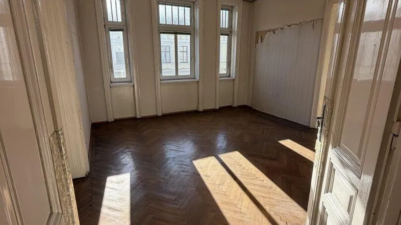 Helles Zimmer mit Fischgrätparkett und hohen Fenstern, durch die Sonnenlicht einfällt.