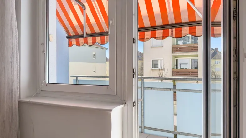 Blick aus dem Fenster auf einen Balkon mit gestreifter Markise und umliegende Gebäude.