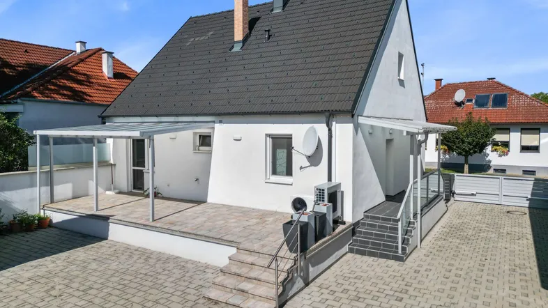 Rückseite des Hauses mit überdachter Terrasse und gepflastertem Bereich, ideal für entspannte Stunden im Freien.