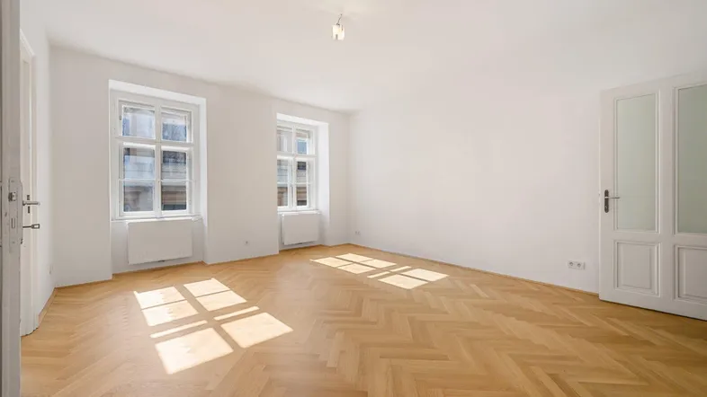Helles Zimmer mit Fischgrätparkettboden und zwei großen Fenstern, durch die Sonnenlicht fällt.