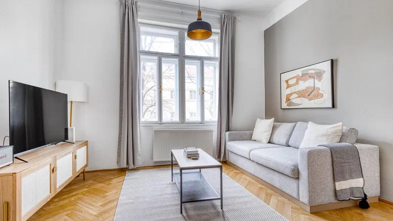 Helles Wohnzimmer mit modernem Sofa, Fernseher und Parkettboden, durch große Fenster gut belichtet.