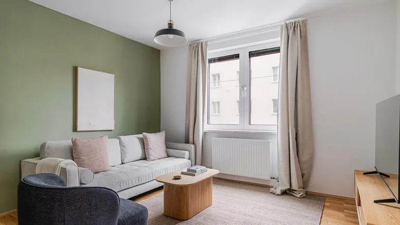 Helles Wohnzimmer mit beiger Couch, grün gestrichener Akzentwand und Parkettboden.