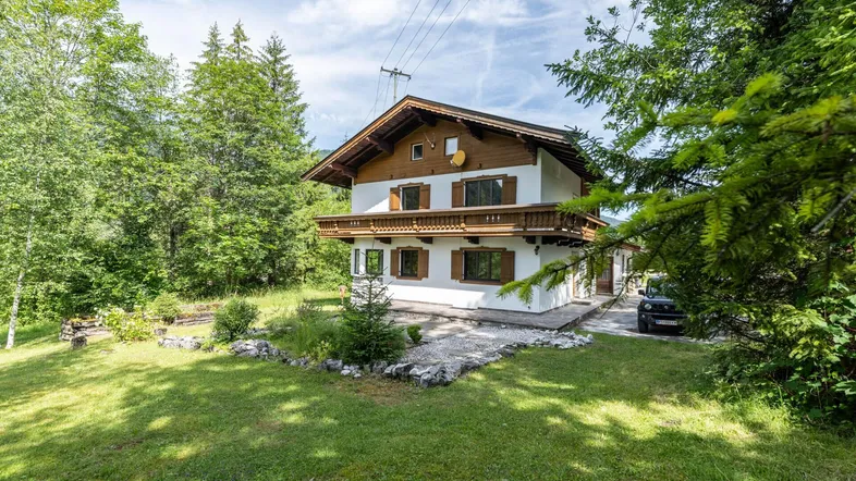 Traditionelles Haus im alpenländischen Stil mit Holzbalkon, umgeben von viel Grün und Bäumen.