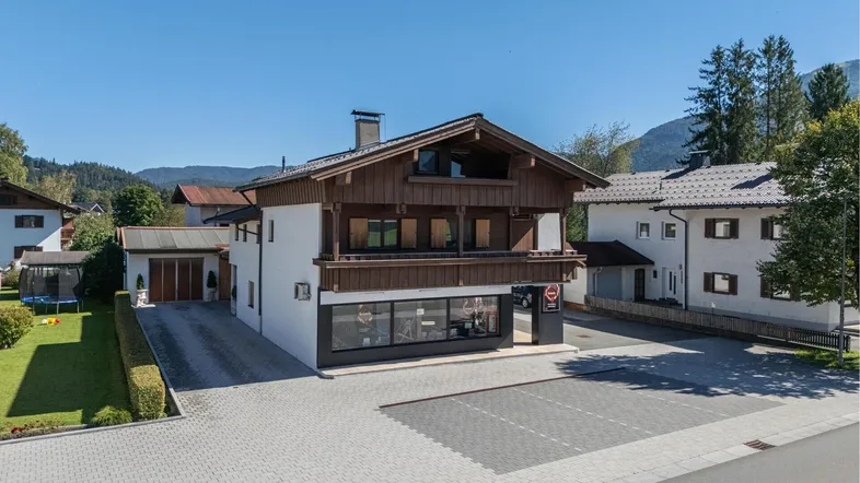 Traditionelles Haus mit Holzfassade, Parkplatz und Bergblick im Hintergrund.