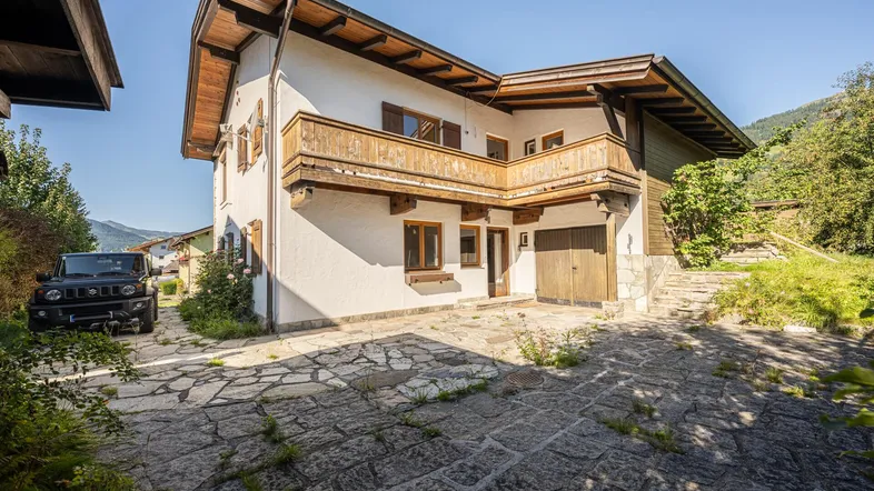 Traditionelles Haus mit Steinfassade, Holzdetails und gepflastertem Vorplatz, umgeben von grüner Natur.