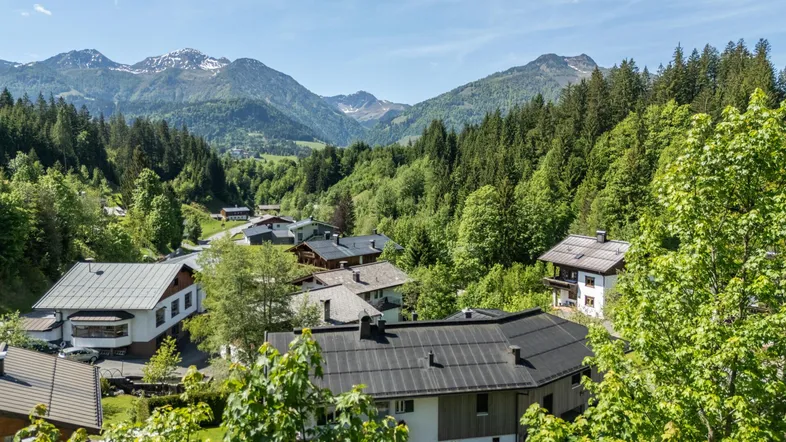 Panoramablick auf ein malerisches Dorf in den Bergen, umgeben von grünen Wäldern und schneebedeckten Gipfeln.