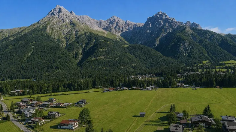 Luftaufnahme eines malerischen Dorfes in den Alpen, umgeben von grünen Wiesen und majestätischen Bergen unter blauem Himmel.