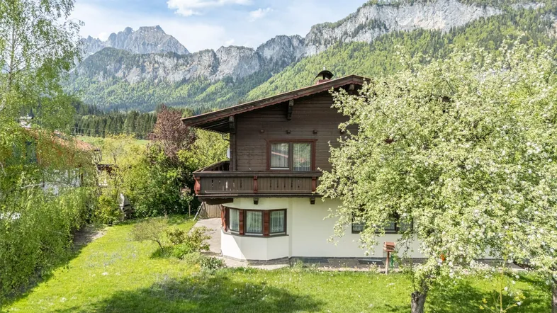 Traditionelles Haus im alpenländischen Stil mit Balkon und Bergblick, umgeben von grüner Natur.