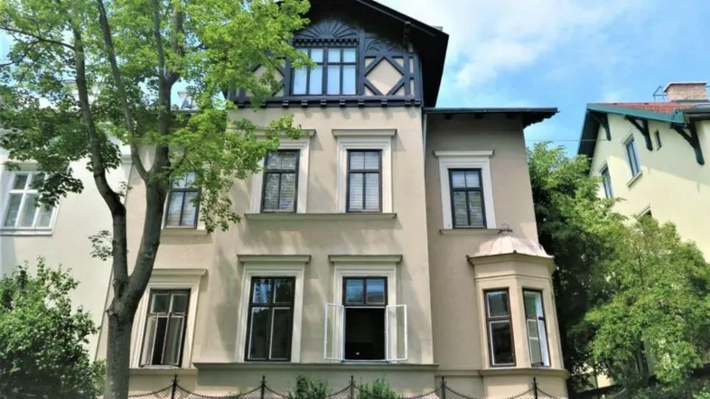 Historisches Gebäude mit klassischer Fassade, großen Fenstern und einem dekorativen Dachgiebel unter blauem Himmel.