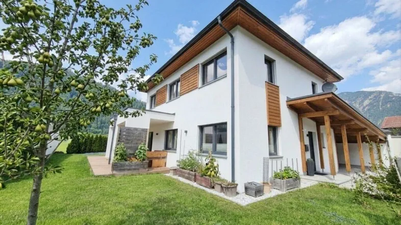 Modernes Einfamilienhaus mit weißer Fassade, Holzdetails und gepflegtem Garten vor Bergkulisse.