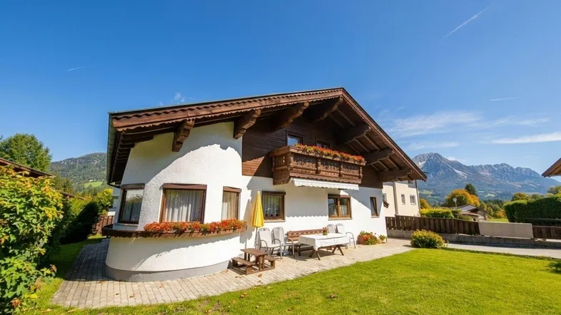 Traditionelles Haus im Alpenstil mit weißer Fassade, Holzbalkonen und Bergblick unter blauem Himmel.