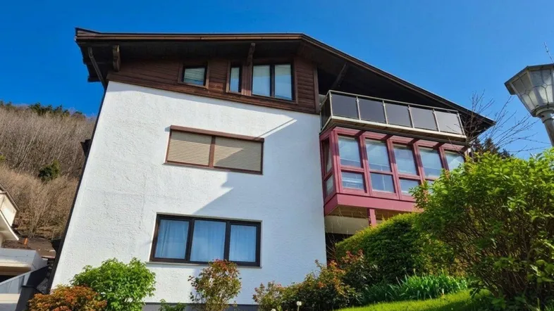 Modernes Haus mit weißer Fassade, Holzverkleidung und einem verglasten Balkon mit Bergblick.