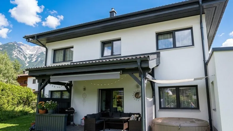 Überdachte Terrasse mit gemütlichem Sitzbereich, Whirlpool und herrlichem Bergblick, umgeben von einem grünen Garten.