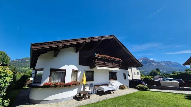 Traditionelles Haus mit Satteldach, Balkon und Garten, umgeben von einer malerischen Berglandschaft unter blauem Himmel.