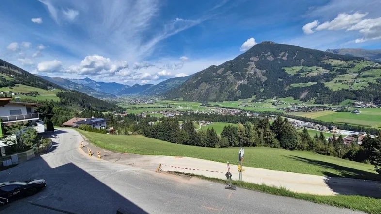 Weitläufiger Blick über ein grünes Tal mit Bergen und einer Straße im Vordergrund unter blauem Himmel.