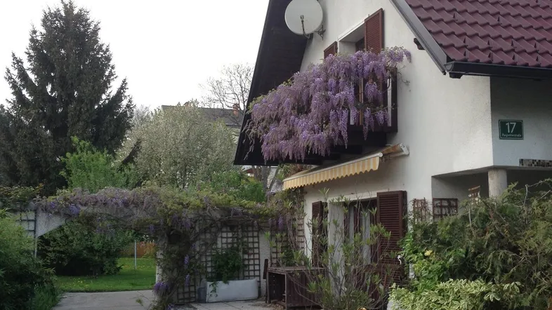 Hausfassade mit blühendem Balkon und einer einladenden Terrasse mit Sitzgelegenheiten im Grünen.