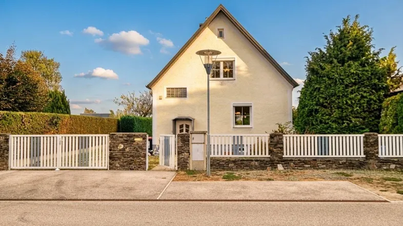 Helles Einfamilienhaus mit Satteldach, weißem Zaun und Steinelementen an der Vorderseite.