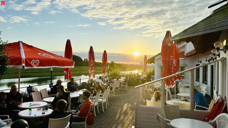 Belebte Außenterrasse mit Gästen, roten Sonnenschirmen und malerischem Sonnenuntergang über dem Fluss.