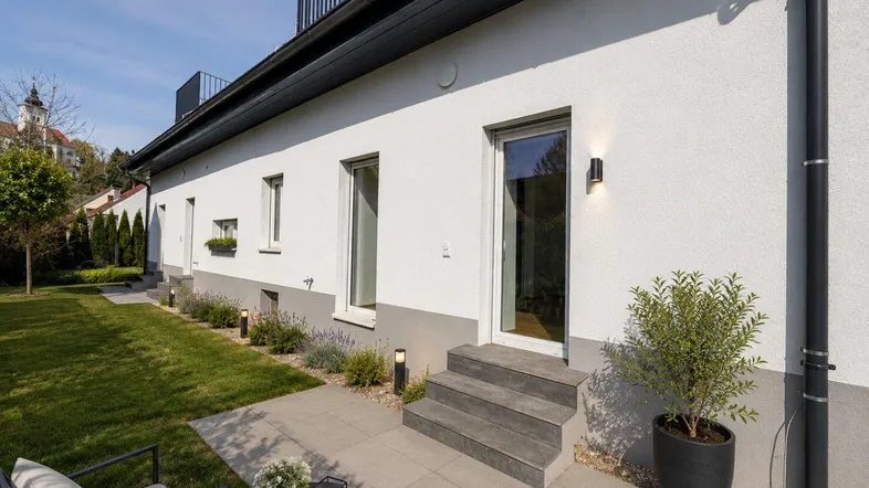 Außenbereich des Hauses mit kleiner Terrasse, Rasenfläche und Zugang zum Gebäude.
