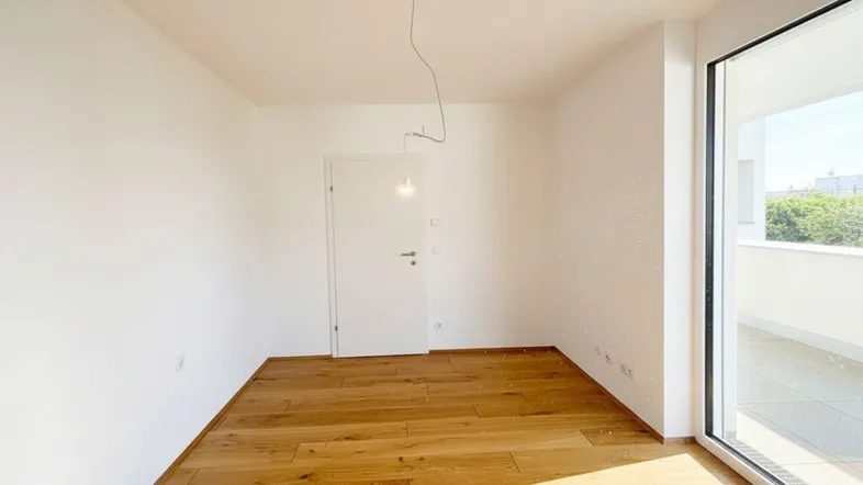 Heller Raum mit Holzboden, weißen Wänden und großem Fenster mit Zugang zum Balkon.