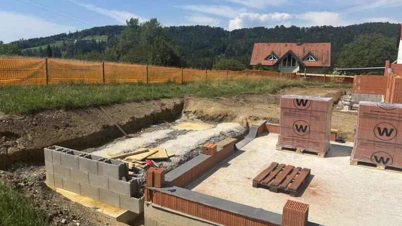 Baustelle mit Fundament und ersten Mauern, im Hintergrund ein Haus mit rotem Dach.