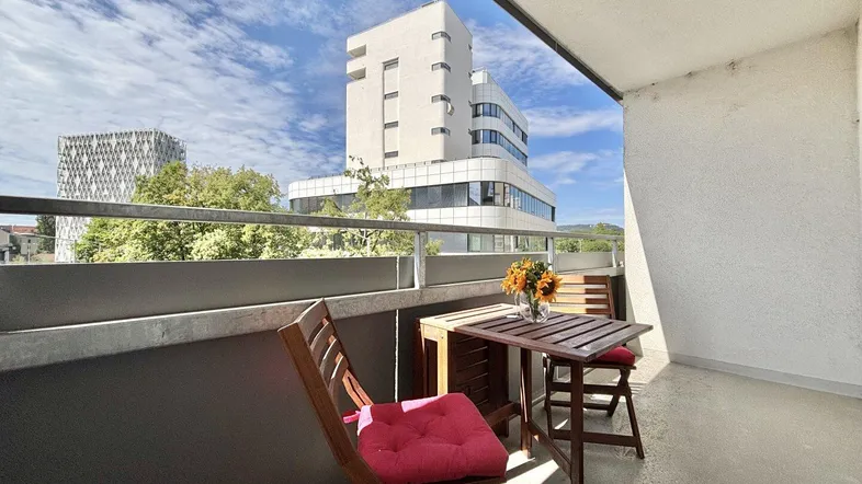 Gemütlicher Balkon mit Holztisch und Stühlen, bietet einen weiten Blick auf moderne Gebäude und den Himmel.