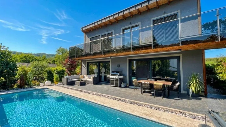 Modernes Haus mit großem Pool, weitläufiger Terrasse und Balkon, umgeben von grüner Landschaft.