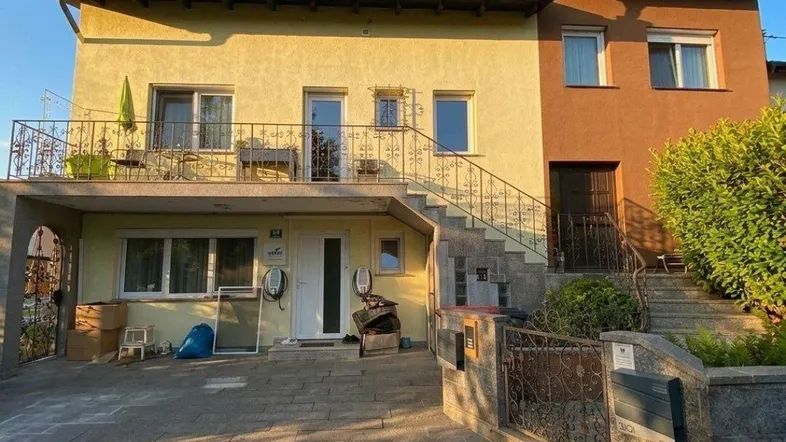 Zweifamilienhaus mit gelber Fassade, Balkon und gepflasterter Auffahrt im Sonnenlicht.
