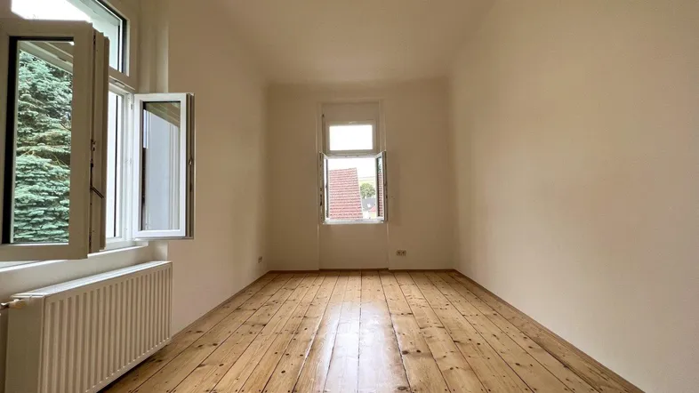 Helles Zimmer mit Holzdielenboden, weißen Wänden und Blick ins Grüne durch das offene Fenster.
