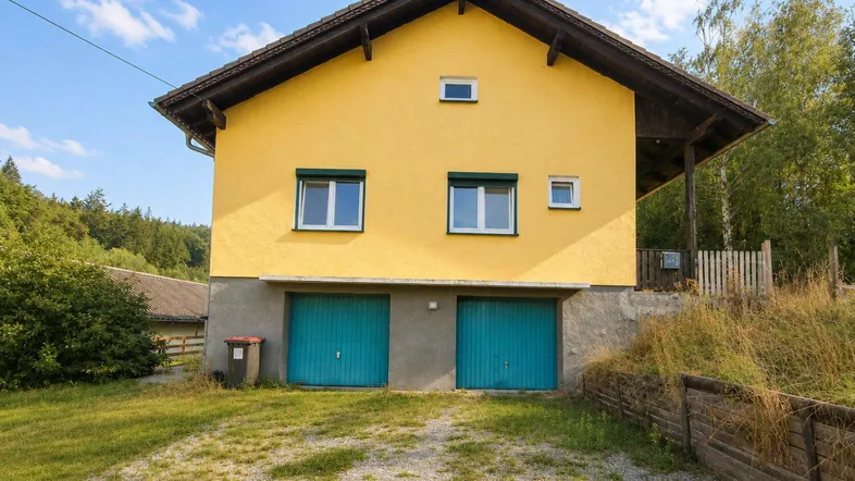 Gelbes Einfamilienhaus mit zwei Garagentoren und umgebender Natur unter blauem Himmel.
