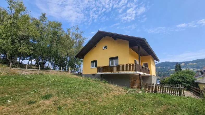 Gelbes Einfamilienhaus mit braunem Dach auf einem grünen Hügel mit Bäumen im Hintergrund.