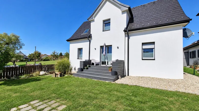 Weißes Einfamilienhaus mit dunklem Dach, kleiner Terrasse und gepflegtem Garten unter blauem Himmel.