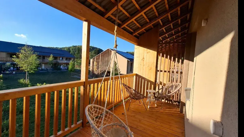 Gemütlicher Holzbalkon mit Hängesessel und Blick auf die umliegende Grünfläche und weitere Gebäude.
