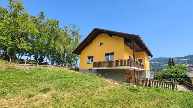 Gelbes Einfamilienhaus auf einem grünen Hügel mit Satteldach und Balkon, umgeben von Bäumen.