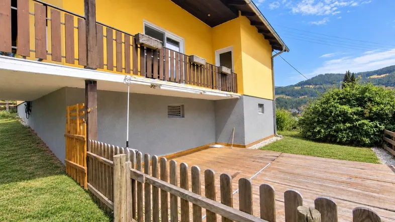 Gelbes Haus mit Holzbalkon und einer Holzterrasse im Gartenbereich mit Blick auf die Landschaft.