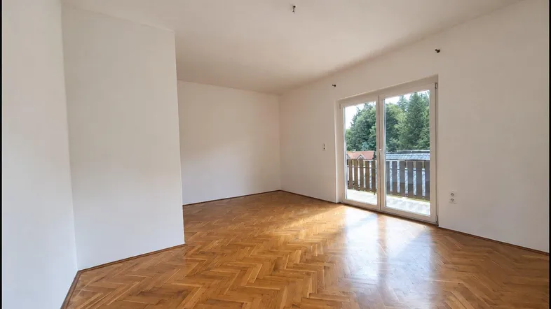 Helles, leeres Wohnzimmer mit Fischgrätparkett und Zugang zu einem Balkon.