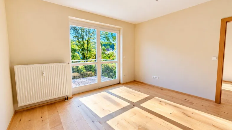 Helles Zimmer mit Holzboden und Zugang zu einem Balkon, bietet viel Tageslicht und einen grünen Ausblick.