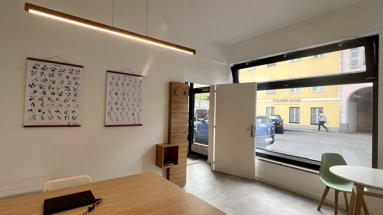 Heller Innenraum mit großem Fenster, Holztisch und Blick auf die Straße, ausgestattet mit einer modernen Deckenleuchte.